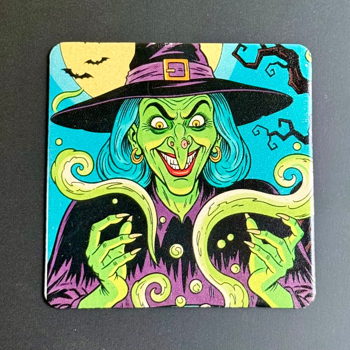 A square coaster featuring a colorful, vintage-style illustration of a witch with green skin, a purple hat and robe, and blue hair, set against a dark gray background.
