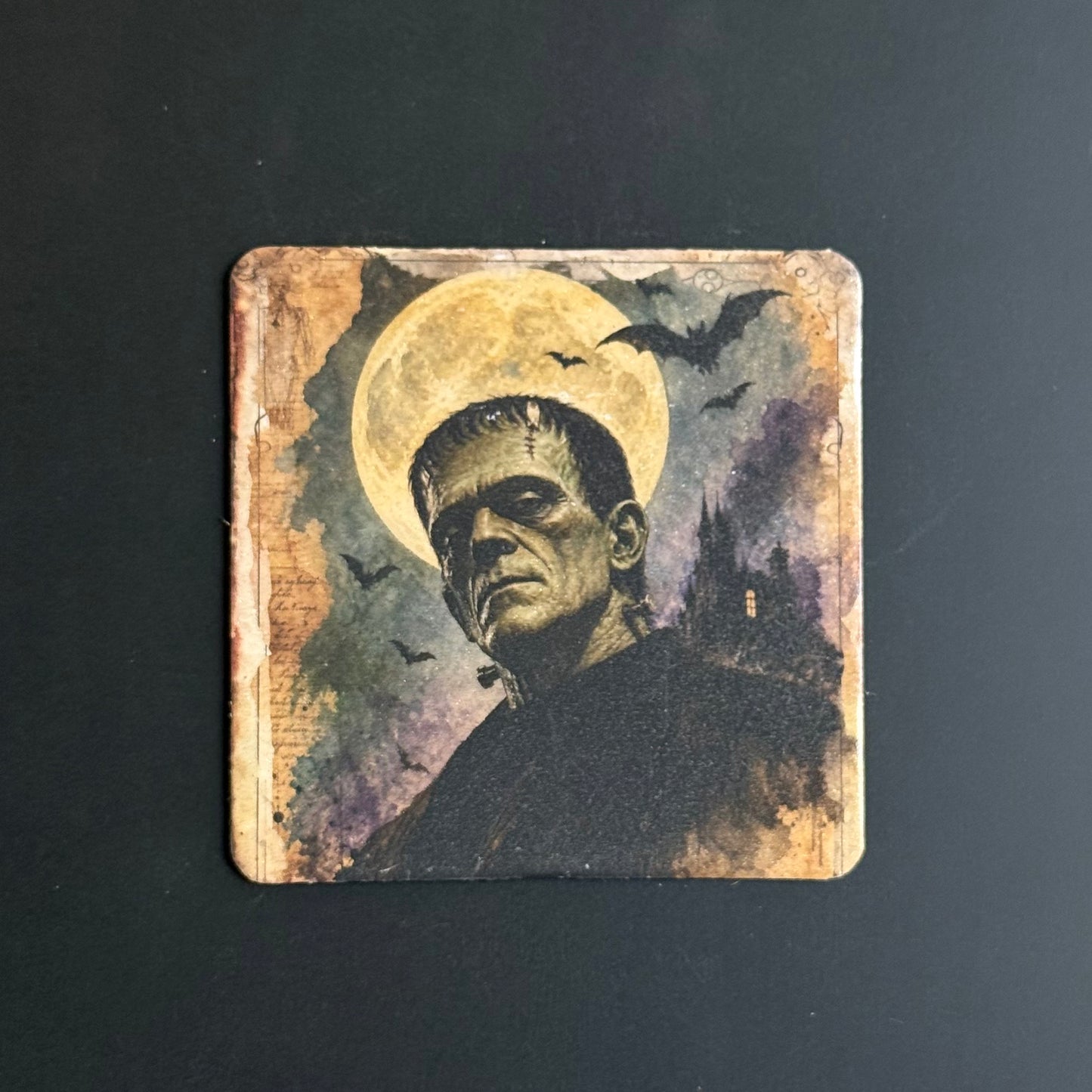 A square coaster featuring a vintage-style painted illustration of Frankenstein's monster in front of a large full moon, with bats and a dark castle in the background, shown on a dark surface.