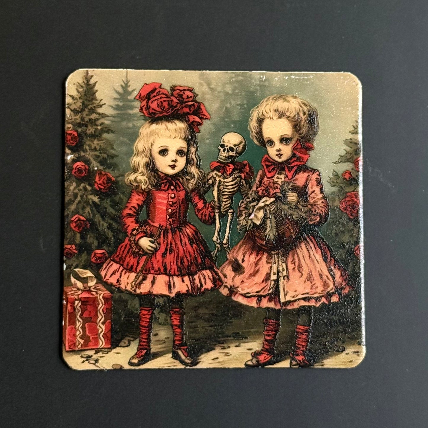 A square coaster featuring a vintage gothic illustration of two young girls in red dresses, one holding a small skeleton and the other standing beside a red gift box, set against a dark forest background.