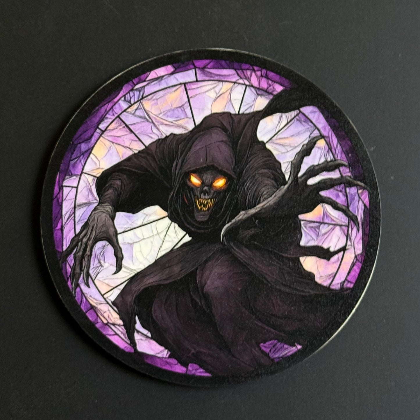 A circular printed coaster or plaque featuring a grim reaper illustration in a stained-glass style, with glowing orange eyes and clawed hands, set against a dark background.
