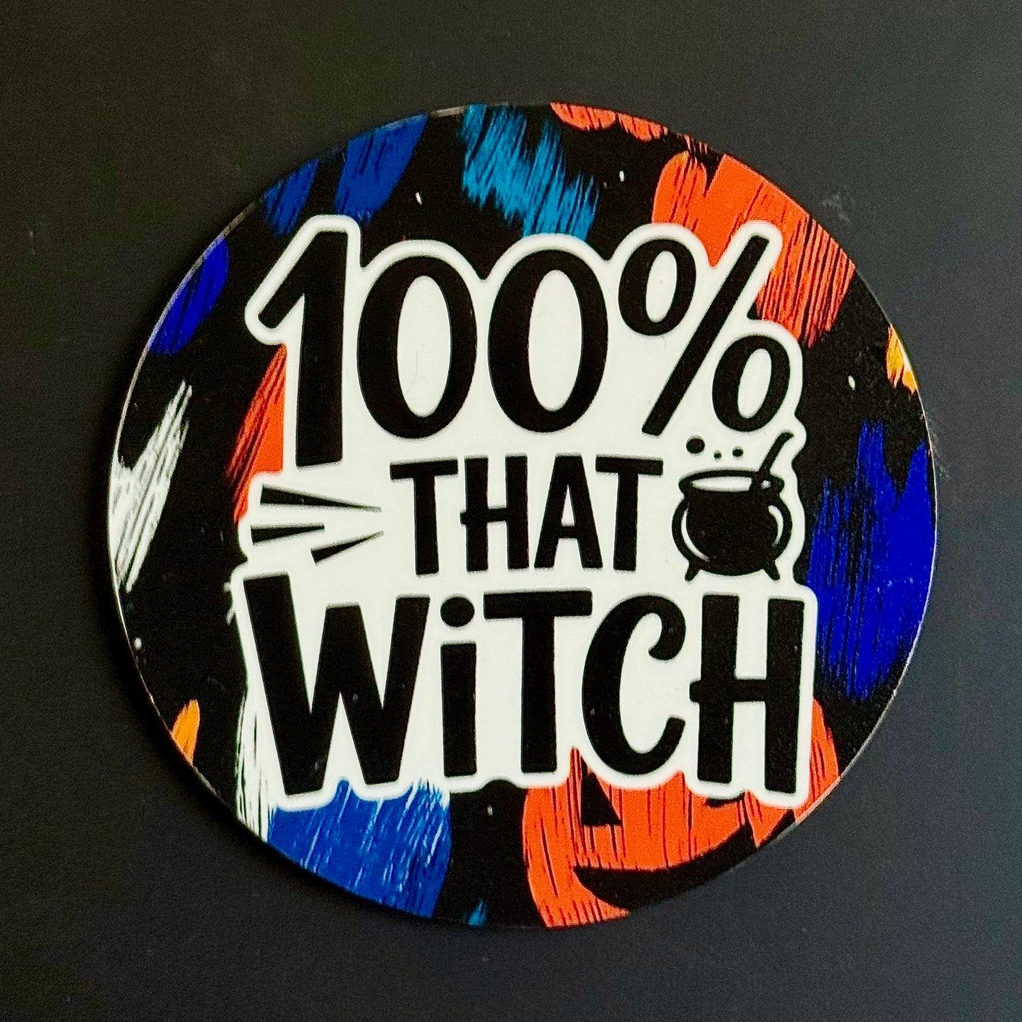 A circular sticker with a Halloween theme featuring the text '100% THAT WITCH' in bold black letters on a white background, surrounded by a colorful abstract pattern of orange, blue, black, and white brushstrokes.