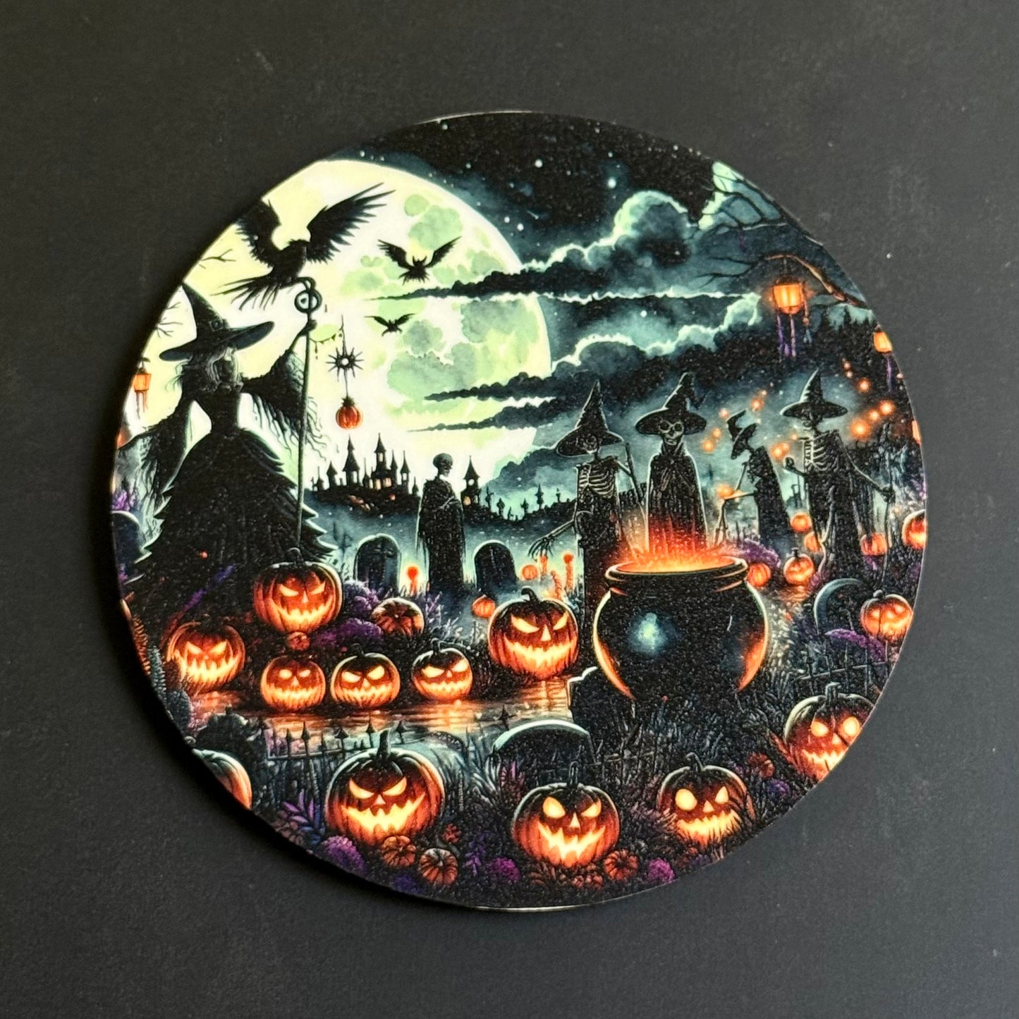 A circular coaster featuring a detailed Halloween night scene with silhouettes of witches, flying bats, a large full moon, haunted houses, glowing jack-o'-lanterns, and a cauldron, photographed on a dark gray background.