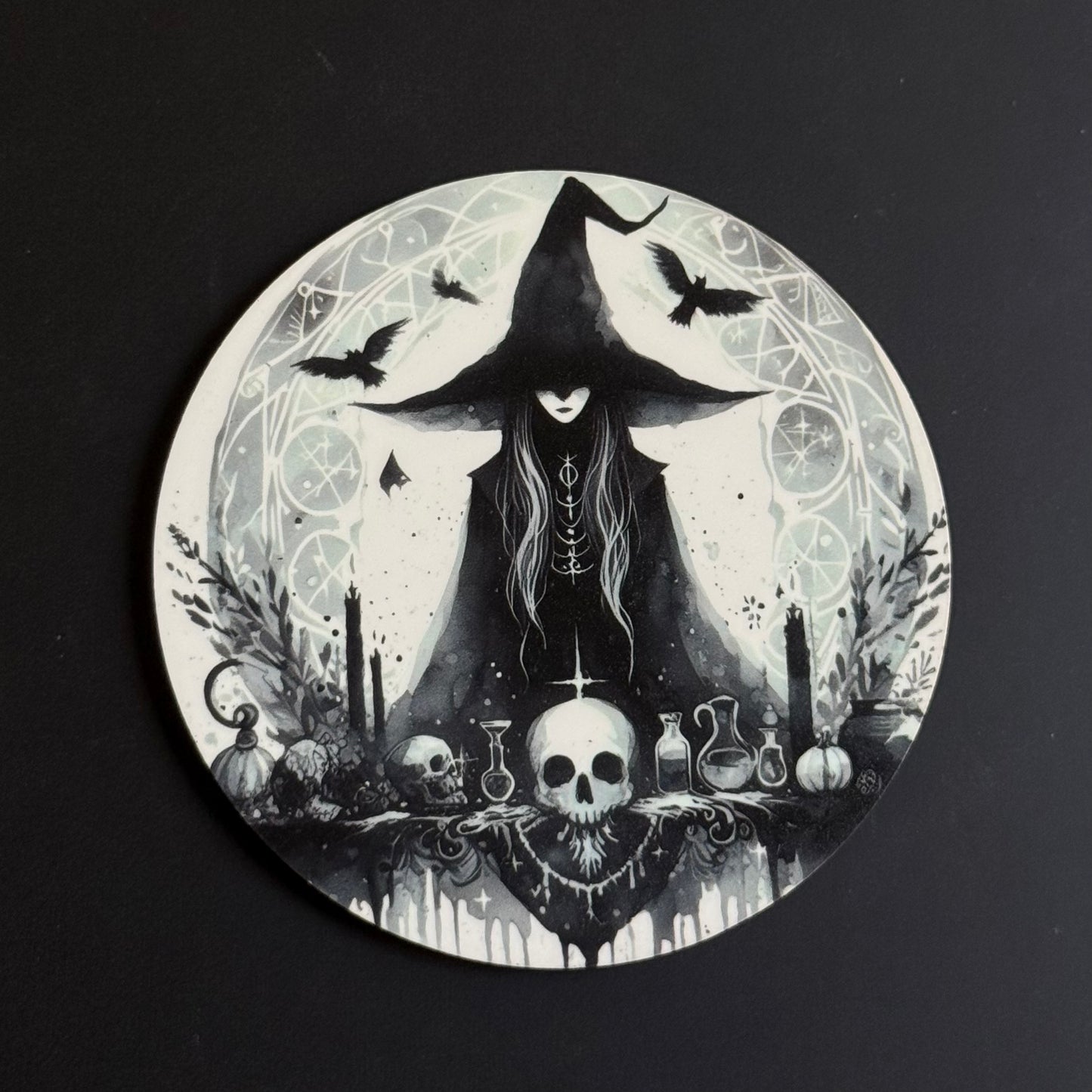 A circular coaster featuring a gothic witch illustration in black and white, depicting a witch in a tall hat with crows, a skull, potions, and candles, set against a dark background.