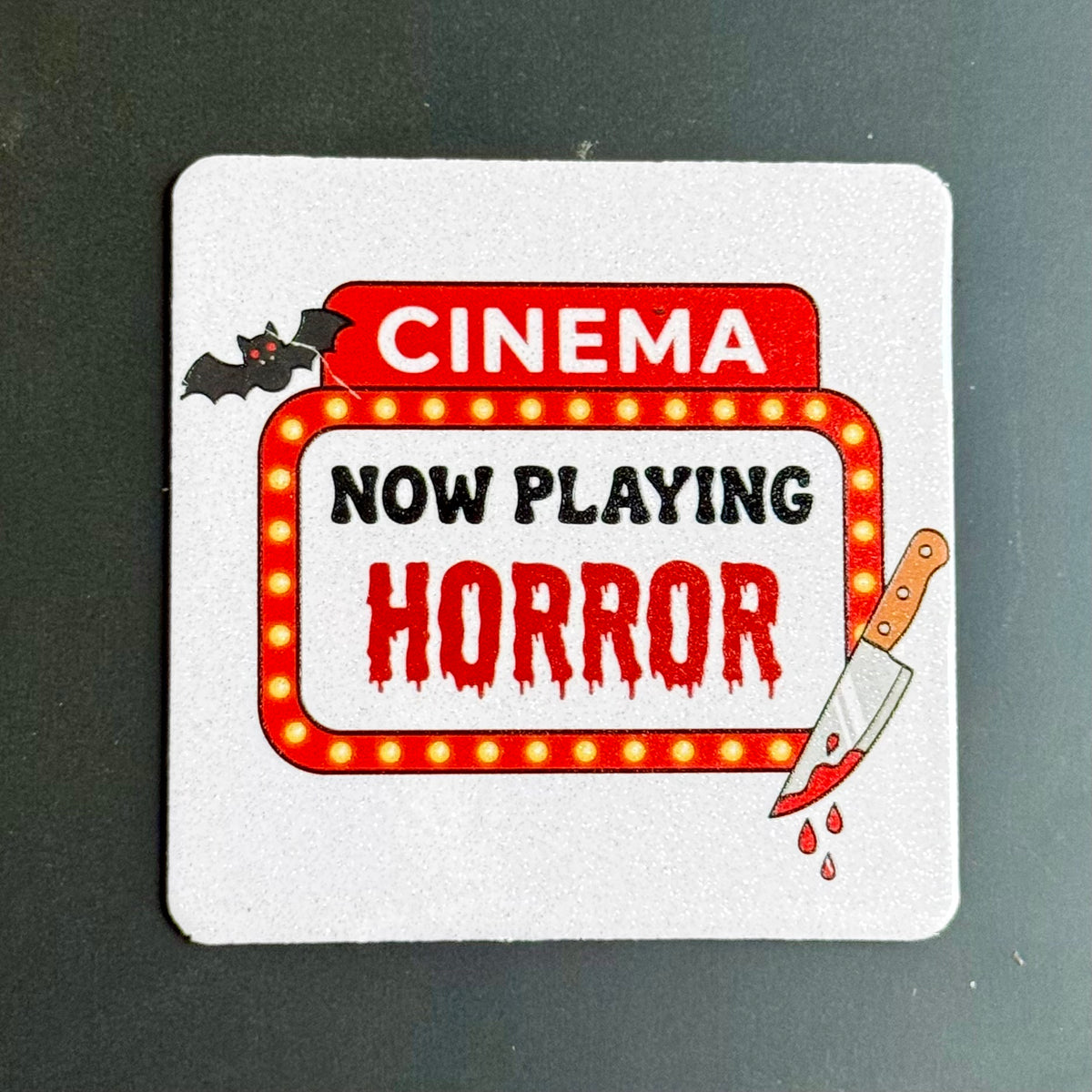 A square white coaster with rounded corners featuring a horror movie theater sign design. The design includes the words 'CINEMA' in white on a red background, 'NOW PLAYING' in black, and 'HORROR' in dripping red letters inside a marquee with yellow light bulbs.
