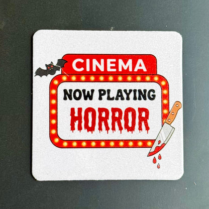 A square white coaster with rounded corners featuring a horror movie theater sign design. The design includes the words 'CINEMA' in white on a red background, 'NOW PLAYING' in black, and 'HORROR' in dripping red letters inside a marquee with yellow light bulbs.