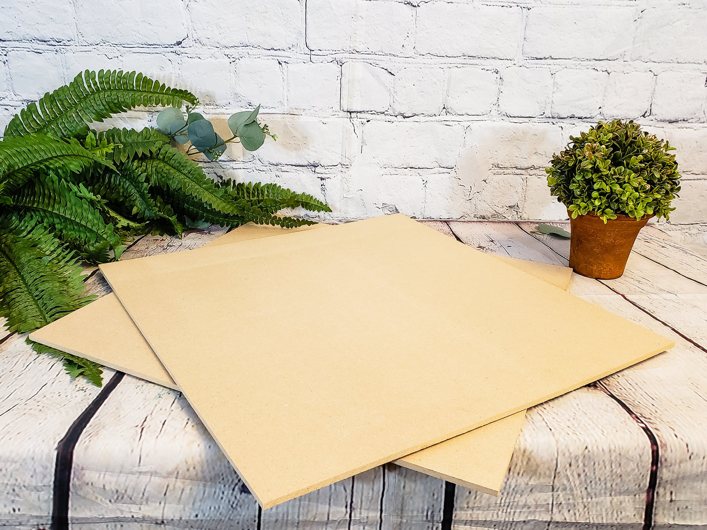 Two beige sheets of MDF on a wooden surface with plants and a white brick wall in the background.