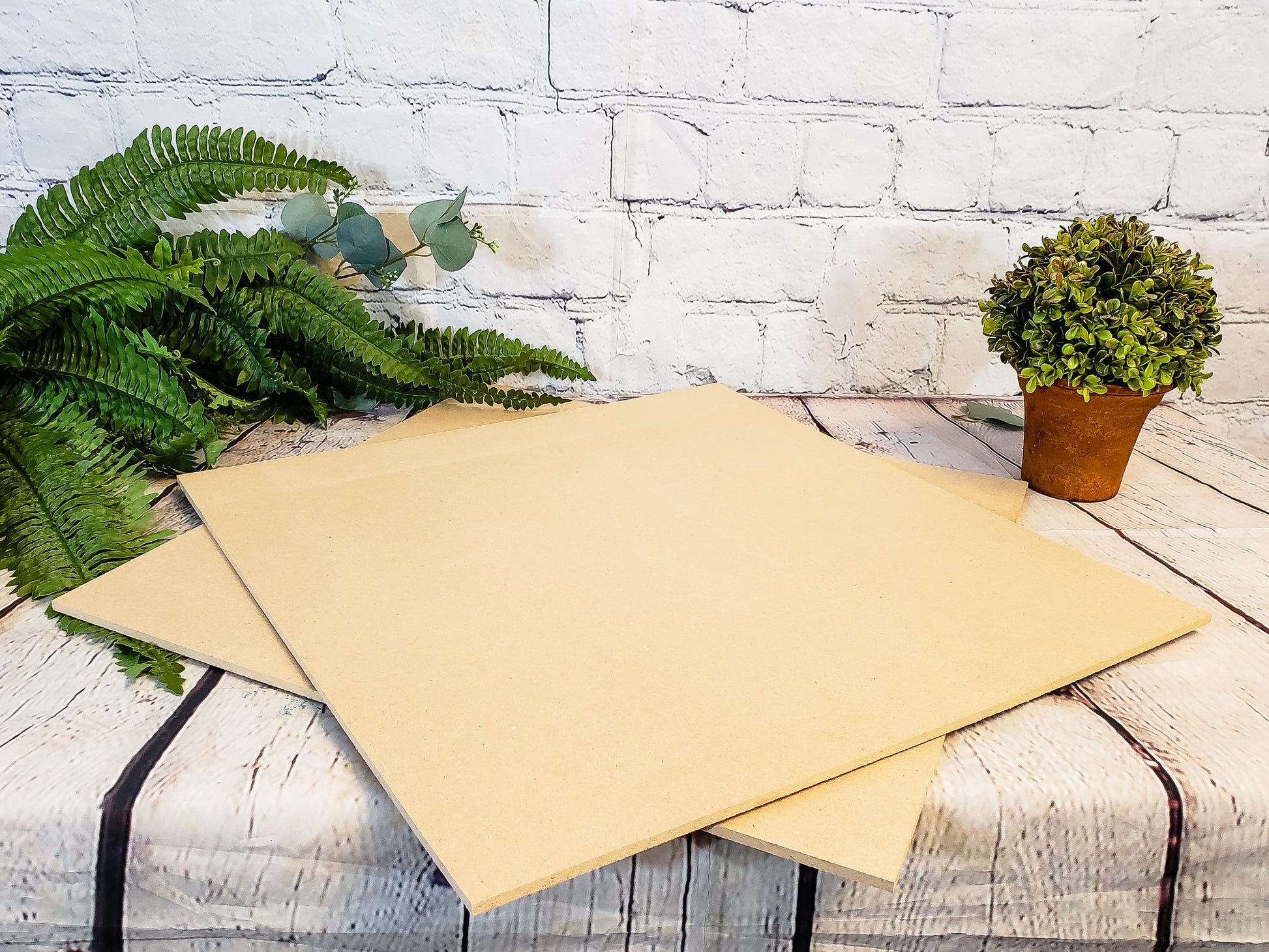 Two beige sheets of MDF on a wooden surface with plants and a white brick wall in the background.