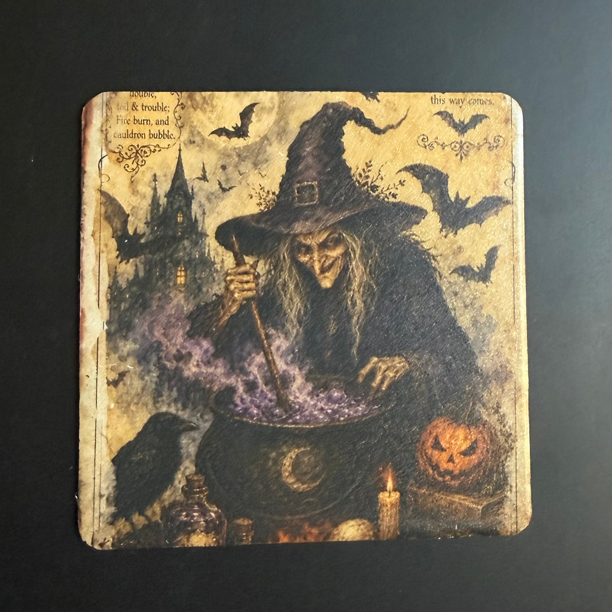 A square coaster featuring a vintage-style Halloween illustration of a witch in a black hat stirring a cauldron with purple smoke, with bats, a haunted house, a pumpkin, and candles on a dark background.