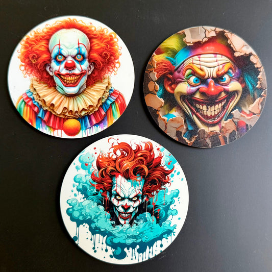 Three round printed coasters featuring horror clown illustrations, arranged on a dark surface. The coasters show colorful clown faces with red hair, blue and orange eye makeup, and dramatic painted backgrounds.