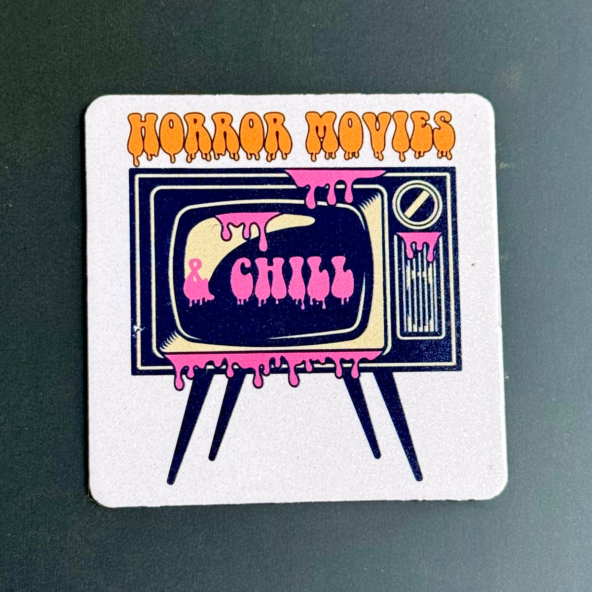 A square white coaster with rounded corners featuring a retro television illustration and the words 'HORROR MOVIES' in orange dripping letters, placed on a dark green surface.