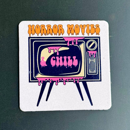 A square white coaster with rounded corners featuring a retro television illustration and the words 'HORROR MOVIES' in orange dripping letters, placed on a dark green surface.