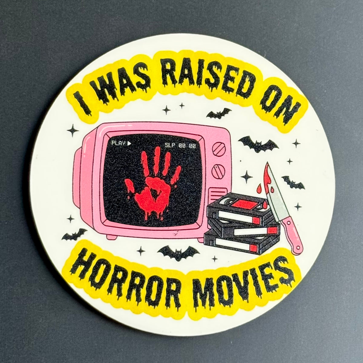 A round sticker or coaster featuring a horror movie theme with the text 'I WAS RAISED ON HORROR MOVIES' in black on a yellow background. The center illustration shows a pink television with a red blood handprint on the screen, a stack of VHS tapes, and a knife, surrounded by black bats and stars on a white circular base, photographed on a dark gray surface.