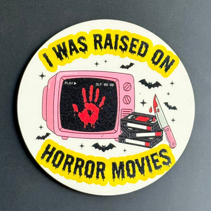 A round sticker or coaster featuring a horror movie theme with the text 'I WAS RAISED ON HORROR MOVIES' in black on a yellow background. The center illustration shows a pink television with a red blood handprint on the screen, a stack of VHS tapes, and a knife, surrounded by black bats and stars on a white circular base, photographed on a dark gray surface.
