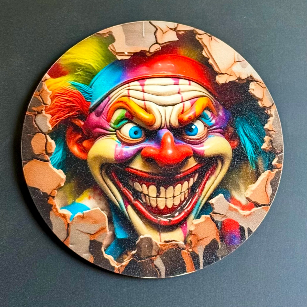 A circular coaster featuring a vibrant, horror-style illustration of a creepy clown bursting through cracked concrete, set against a dark gray background.