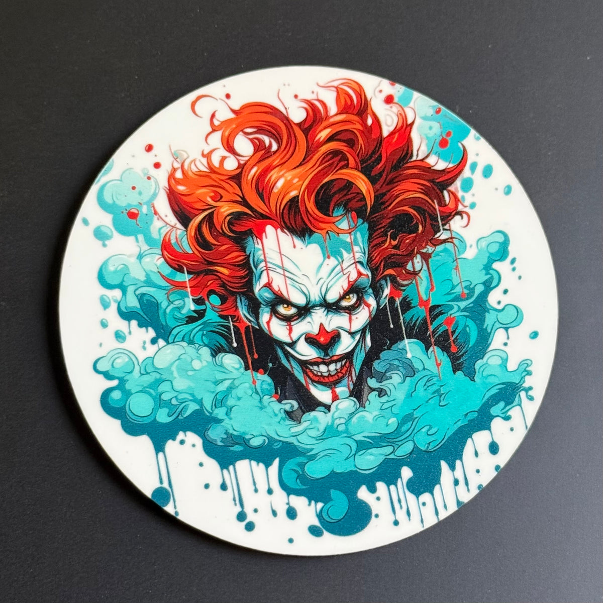 A circular coaster featuring a stylized illustration of a clown with bright red hair, white face paint, and red blood splatters emerging from a swirling teal splash, shown on a dark gray background.