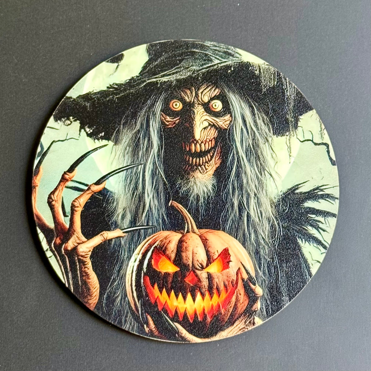 A circular coaster featuring a spooky Halloween illustration of a witch with long gray hair holding a glowing jack-o'-lantern pumpkin, shown on a dark gray background.
