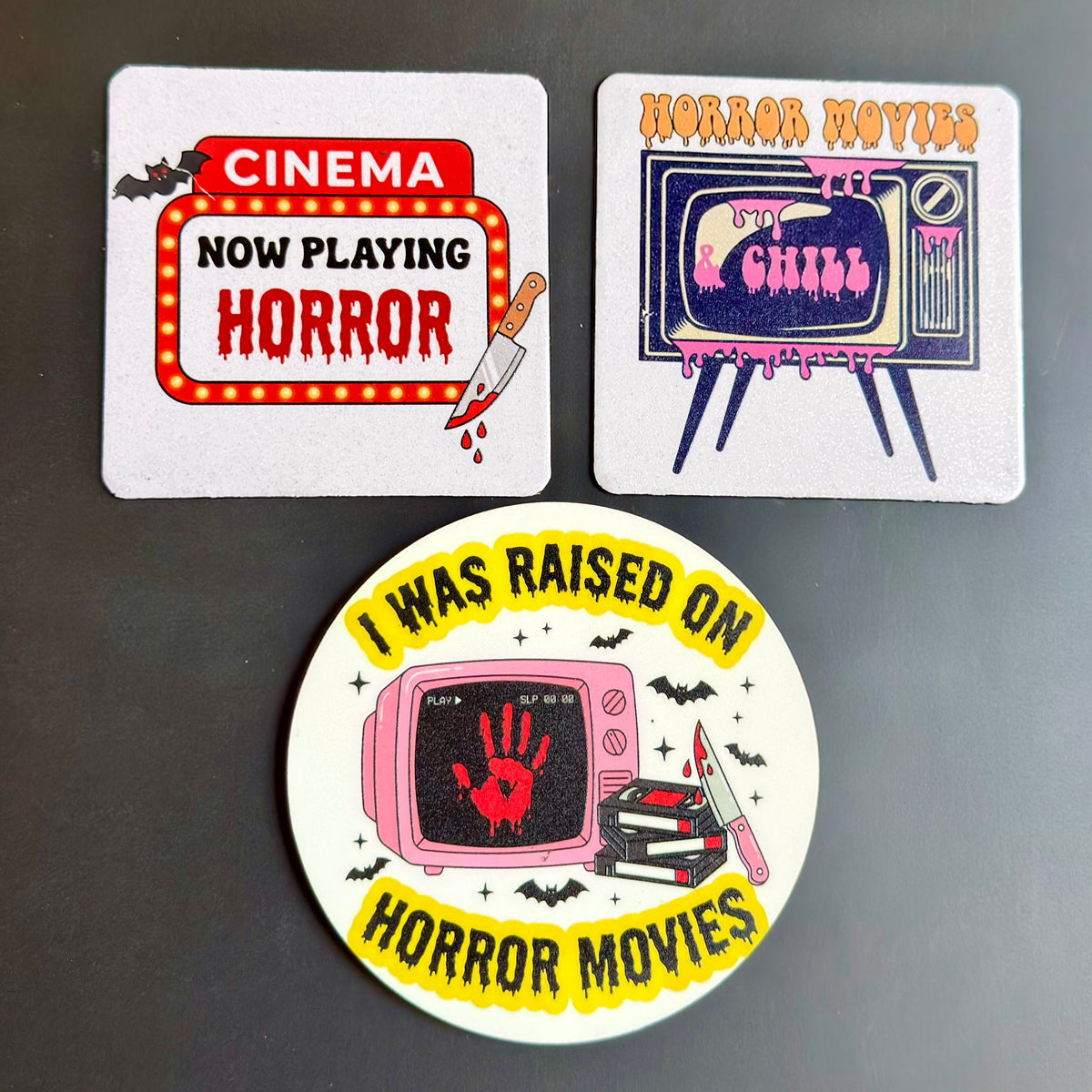 Three horror-themed printed items on a dark gray background: a square coaster reading 'CINEMA NOW PLAYING HORROR,' a square card reading 'HORROR MOVIES' with a retro TV and 'Don't Breathe,' and a round sticker with the text 'I WAS RAISED ON HORROR MOVIES' around a pink TV with a red handprint.