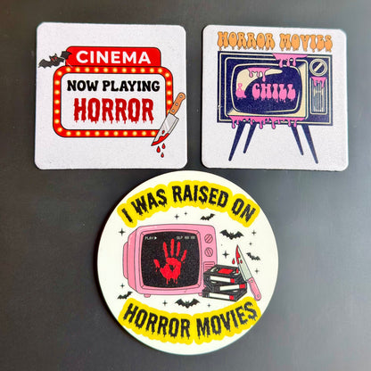 Three horror-themed printed items on a dark gray background: a square coaster reading 'CINEMA NOW PLAYING HORROR,' a square card reading 'HORROR MOVIES' with a retro TV and 'Don't Breathe,' and a round sticker with the text 'I WAS RAISED ON HORROR MOVIES' around a pink TV with a red handprint.