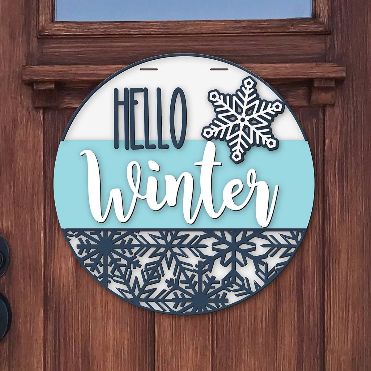 Round door hanger reading Hello Winter in navy and white on an ice blue band, with a raised snowflake and a navy snowflake pattern along the bottom, hanging on a wooden front door.