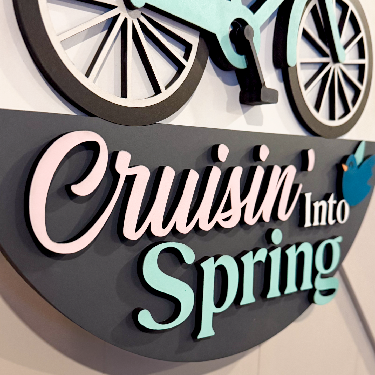 Decorative sign with bicycle design and 'Cruisin' Into Spring' text on a white background