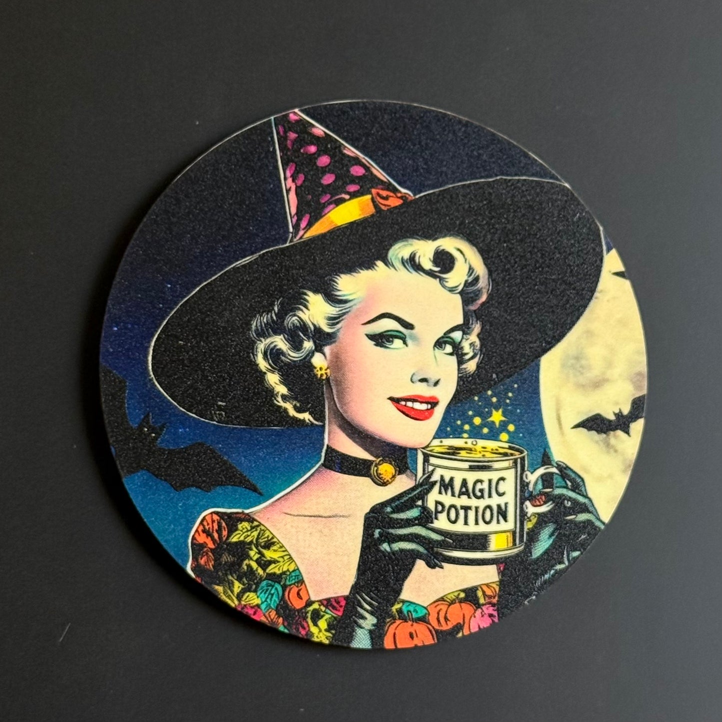 A round printed coaster or plaque featuring a retro comic-style illustration of a woman in a witch hat holding a can labeled 'MAGIC POTION,' set against a dark background with bats and a moon.