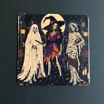 A square Halloween-themed coaster featuring three skeleton figures: a ghost in white, a witch in purple and orange holding a broom, and a mummy wrapped in bandages, set against a dark background with a full moon and haunted houses.