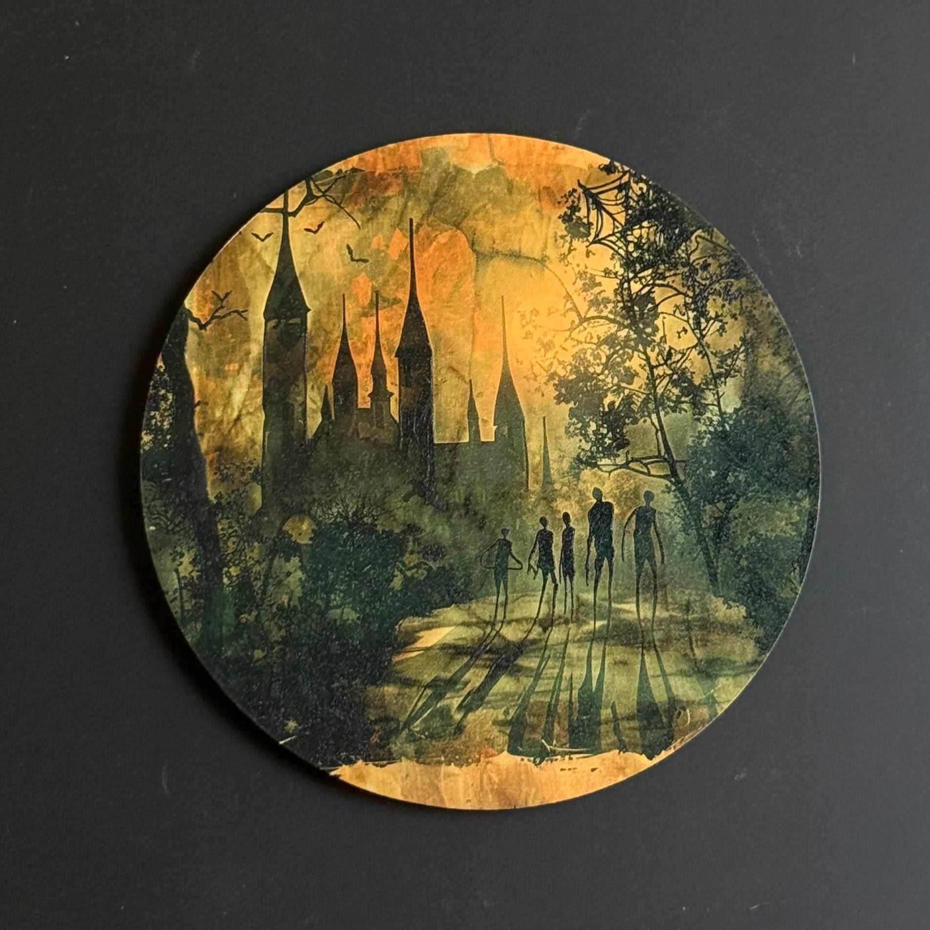 A circular coaster featuring a gothic fantasy illustration of a castle in a misty forest at night, with silhouetted figures walking along a path and a warm orange glow, set against a dark gray background.