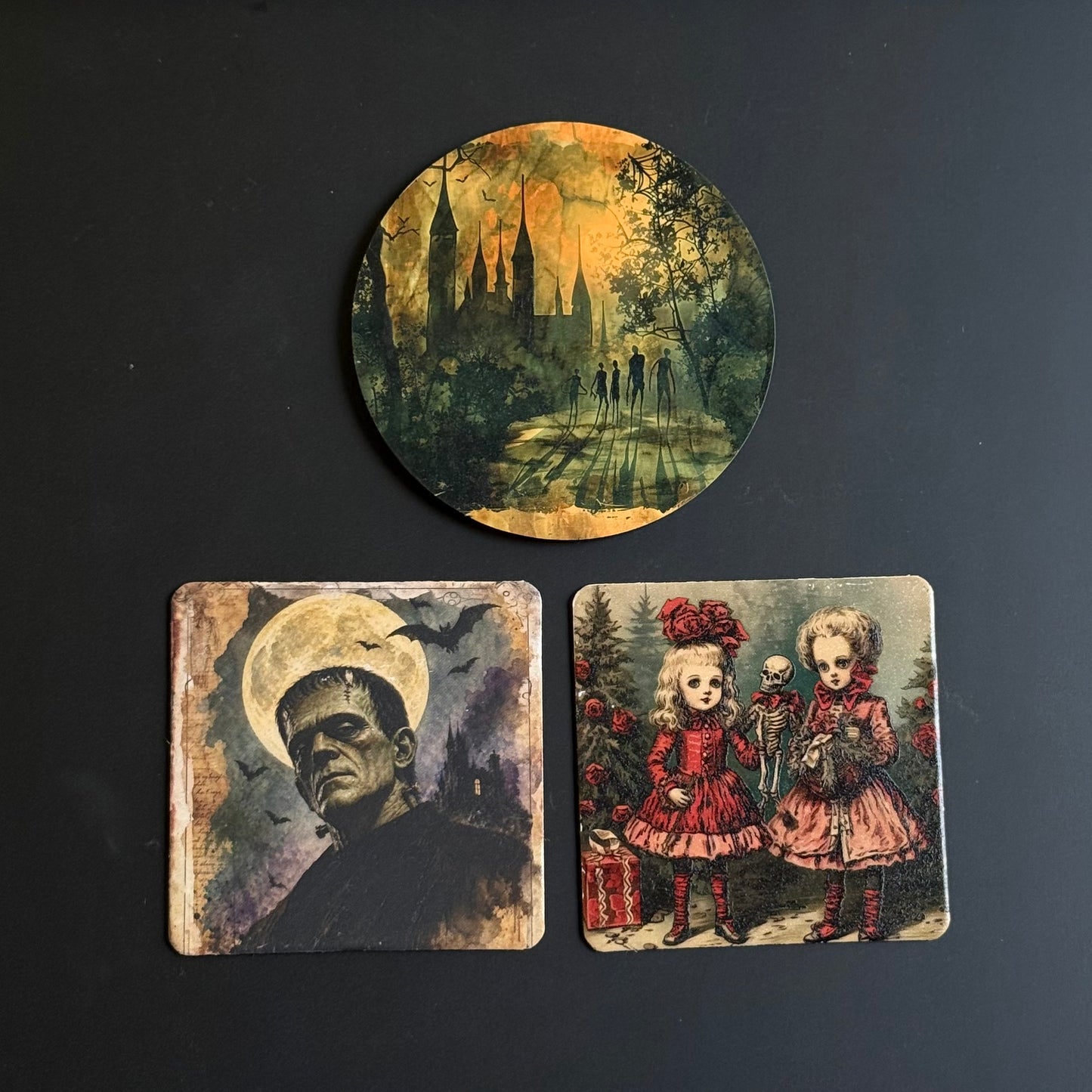 Three printed coasters displayed on a dark background: one round coaster above shows a moody castle scene in green, yellow, and brown tones, and two square coasters below feature vintage horror illustrations, including Frankenstein and two young girls in red dresses.