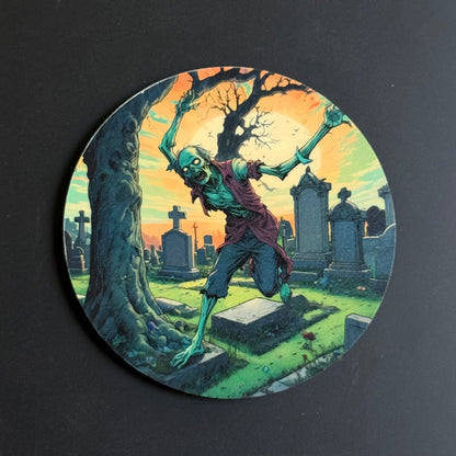 A circular coaster featuring a detailed illustration of a zombie in a spooky graveyard at sunset, photographed on a dark gray background.