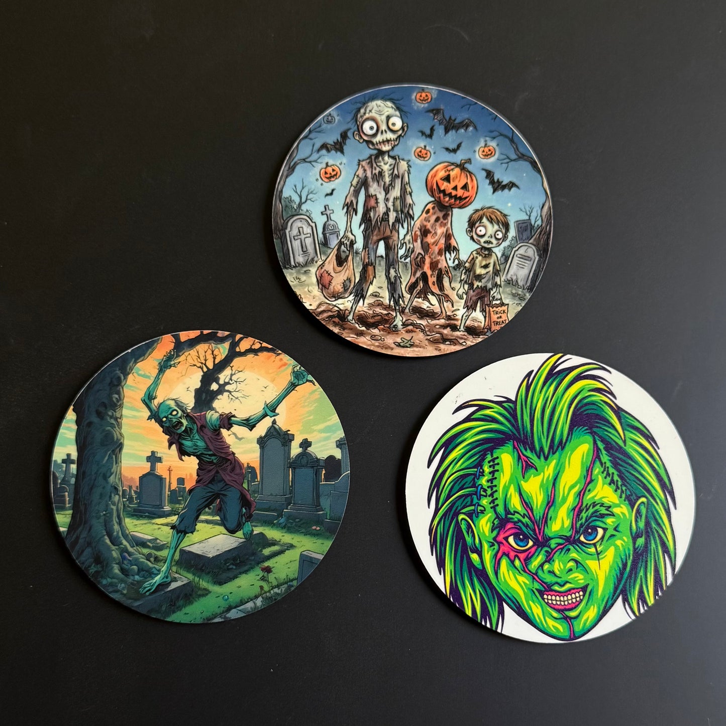 Three circular printed coasters with Halloween-themed illustrations, arranged on a dark background. One coaster shows a zombie, a child, and a pumpkin in a graveyard; another shows a zombie in a cemetery at sunset; the third features a green horror character face on a white background.
