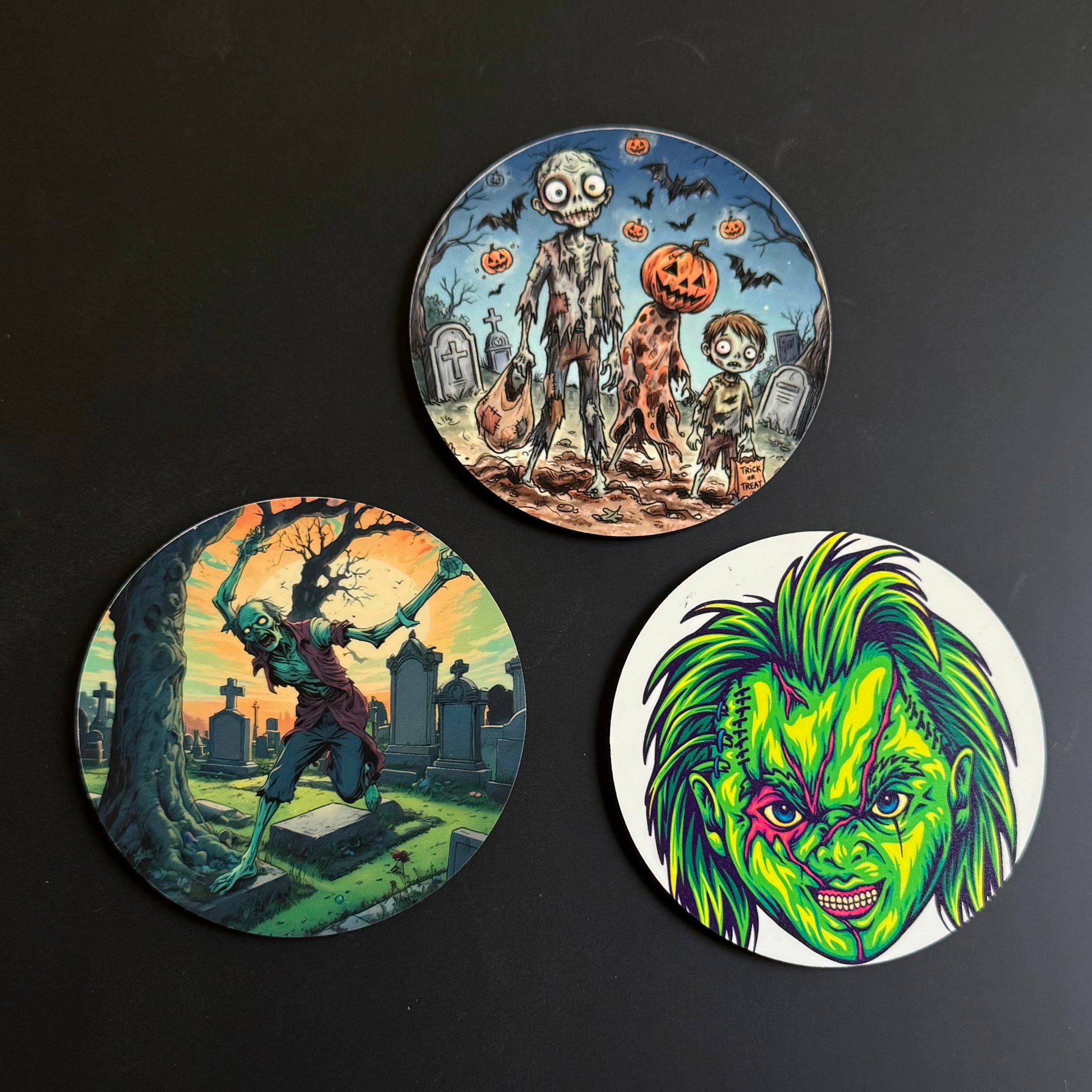 Three circular printed coasters with Halloween-themed illustrations, arranged on a dark background. One coaster shows a zombie, a child, and a pumpkin in a graveyard; another shows a zombie in a cemetery at sunset; the third features a green horror character face on a white background.