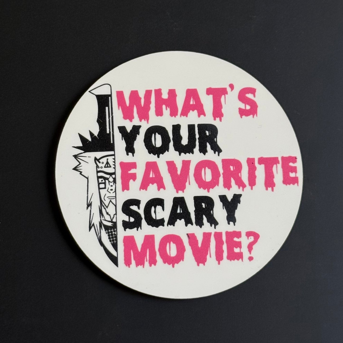A round white sticker or coaster featuring the text 'WHAT'S YOUR FAVORITE SCARY MOVIE?' in dripping pink and black lettering, alongside a black-and-white illustrated knife, shown on a dark background.