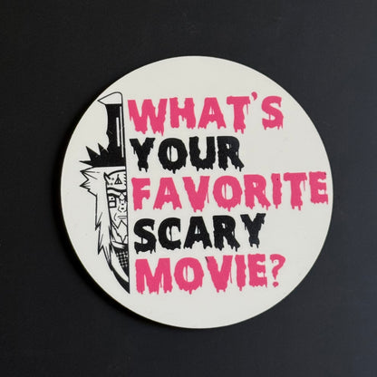 A round white sticker or coaster featuring the text 'WHAT'S YOUR FAVORITE SCARY MOVIE?' in dripping pink and black lettering, alongside a black-and-white illustrated knife, shown on a dark background.