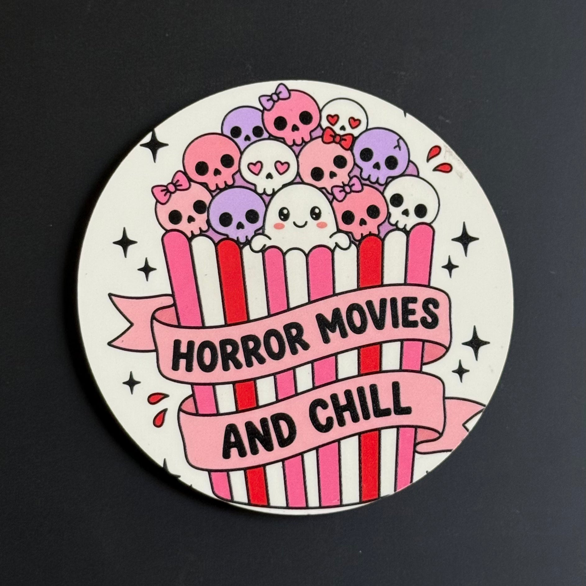 A round sticker or coaster featuring a cartoon illustration of skulls and a ghost popping out of a pink-and-white striped popcorn bucket, with a banner reading 'HORROR MOVIES AND CHILL' on a dark background.