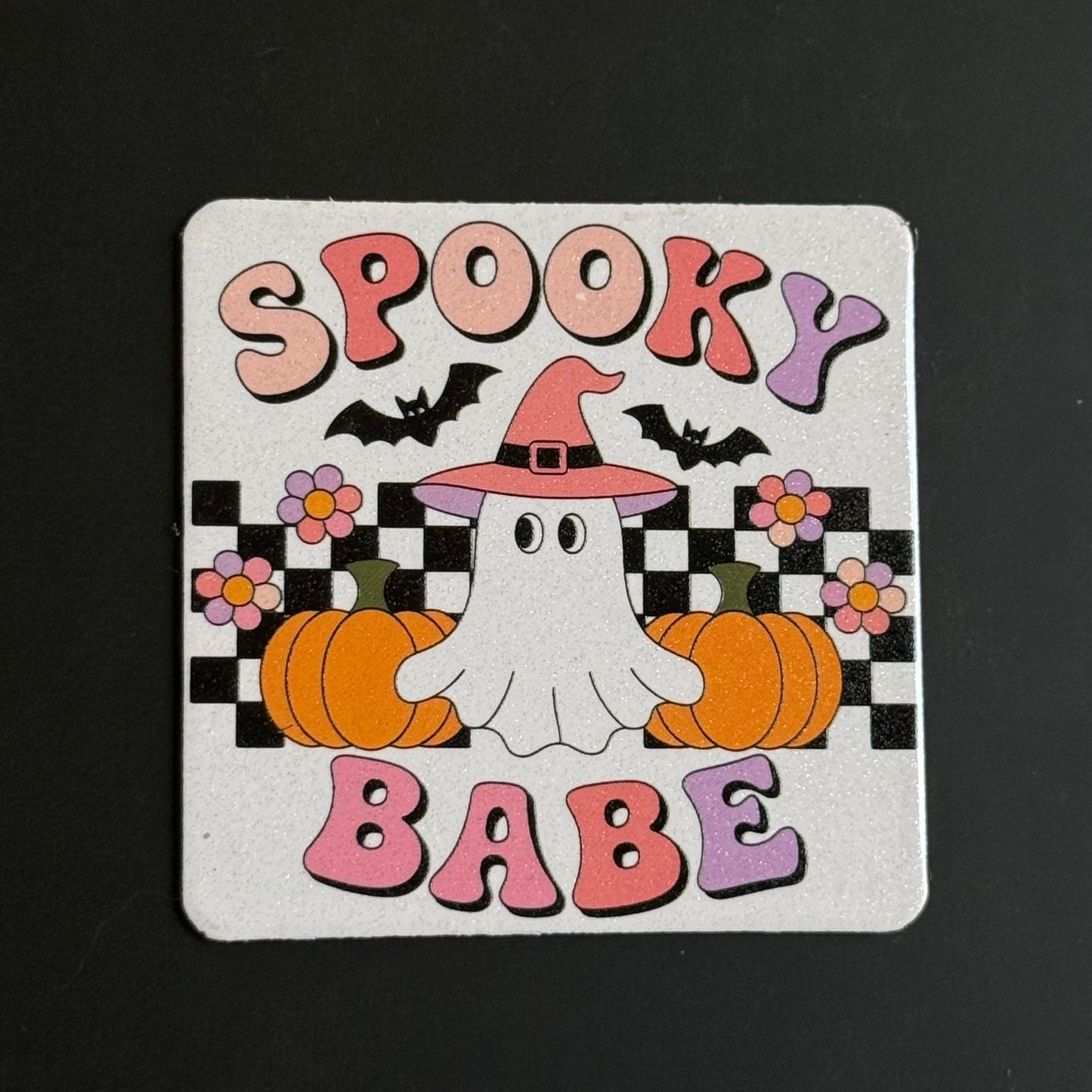 A square white coaster with rounded corners featuring a colorful Halloween design. The design includes the words 'SPOOKY BABE' in pink and purple bubble letters, two black bats, a white cartoon ghost wearing a pink witch hat, two orange pumpkins, and pink and purple flowers over a black and white checkered background.