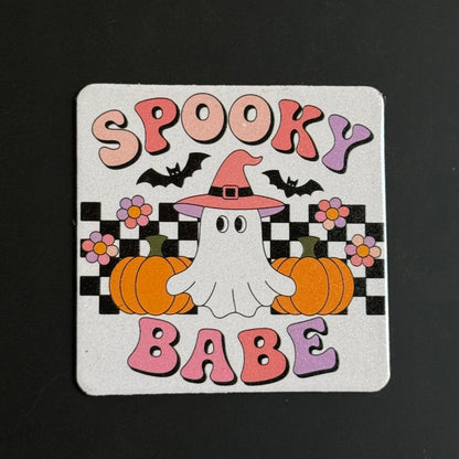 A square white coaster with rounded corners featuring a colorful Halloween design. The design includes the words 'SPOOKY BABE' in pink and purple bubble letters, two black bats, a white cartoon ghost wearing a pink witch hat, two orange pumpkins, and pink and purple flowers over a black and white checkered background.