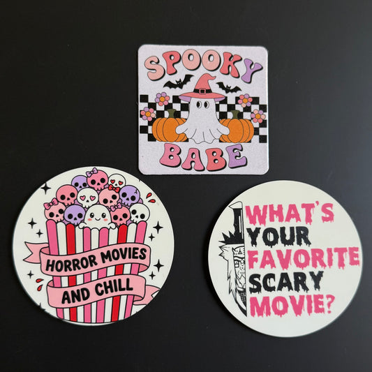 Three Halloween-themed printed stickers or badges arranged on a dark background: a square card reading “Spooky Babe” with a ghost, pumpkins, bats, and flowers, and two round badges with horror movie text and skull artwork.