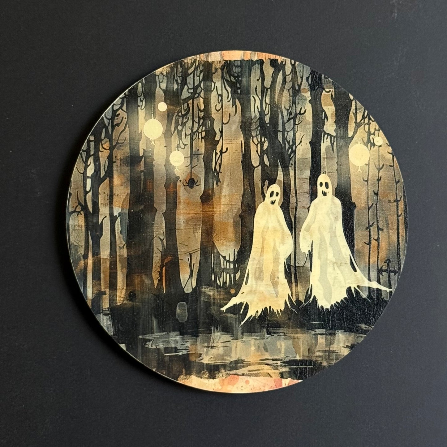 A circular art print or coaster featuring a spooky forest scene with two white ghost figures among dark, bare trees under a warm brown and beige sky, photographed against a dark background.