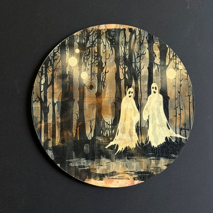 A circular art print or coaster featuring a spooky forest scene with two white ghost figures among dark, bare trees under a warm brown and beige sky, photographed against a dark background.
