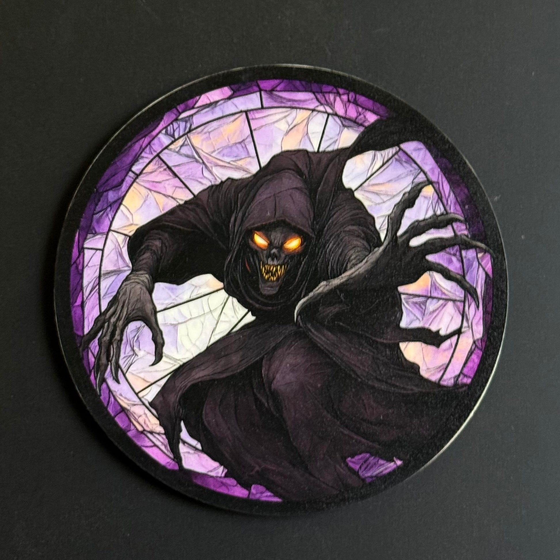 A circular printed coaster or plaque featuring a grim reaper illustration in a stained-glass style, with glowing orange eyes and clawed hands, set against a dark background.