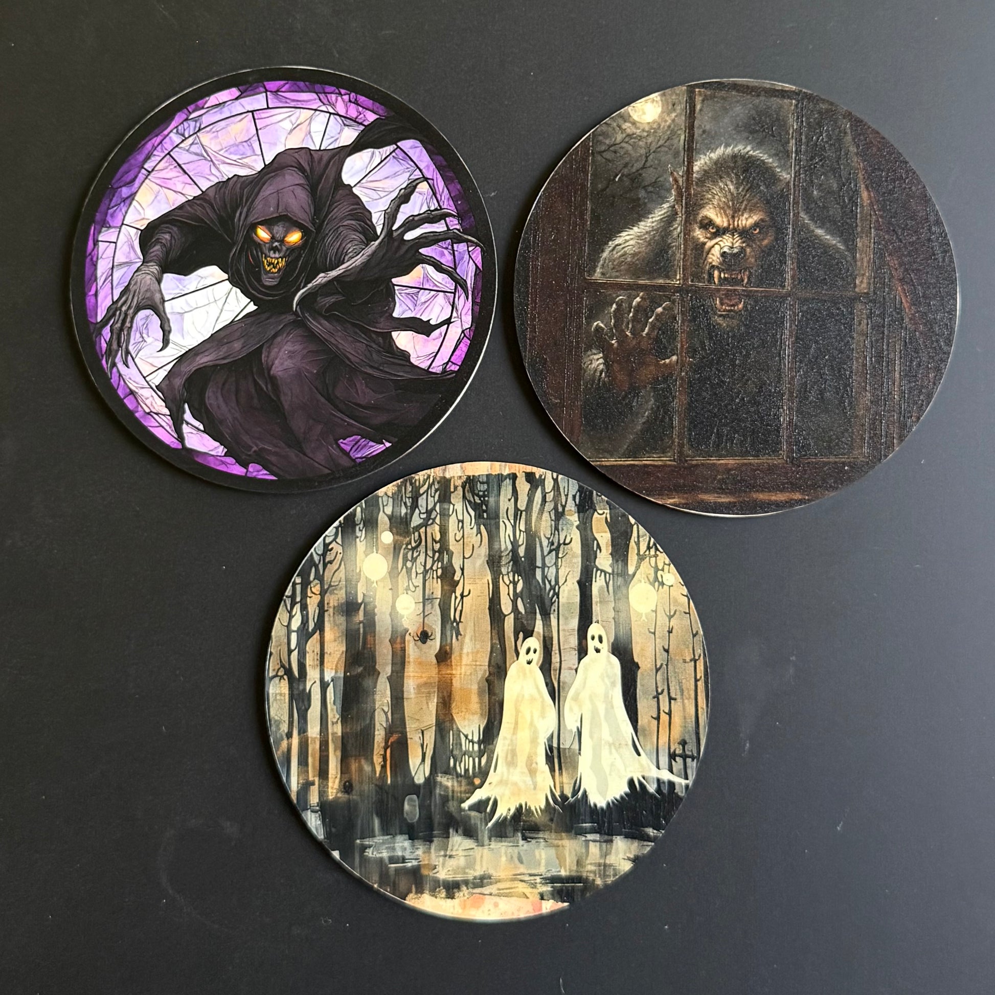 Three round printed coasters with Halloween-themed illustrations, displayed on a dark surface. One coaster shows a hooded figure in a stained-glass style with purple and black tones; another shows a dark ape behind bars in brown and black; the third shows two white ghosts in a spooky forest with beige, black, and white tones.