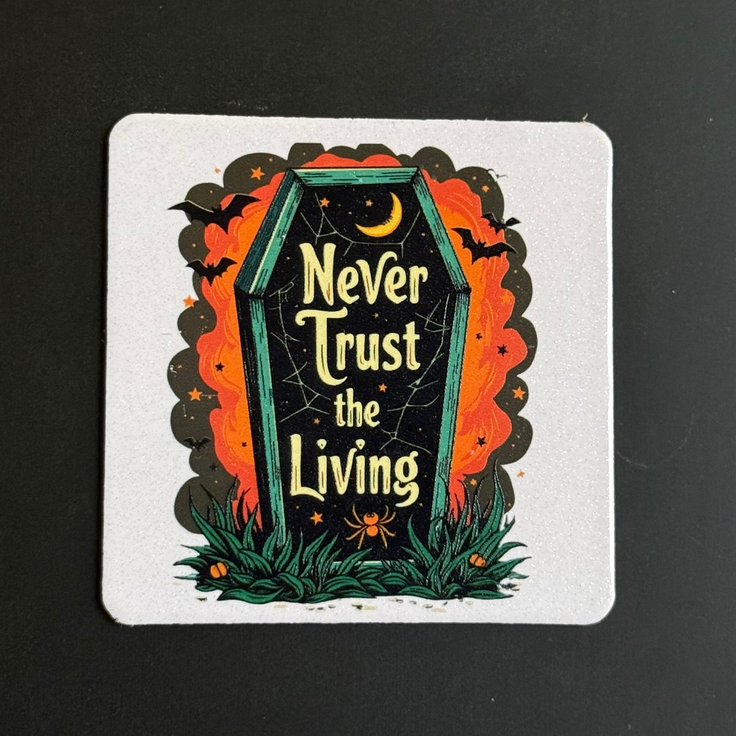 A square white coaster featuring a Halloween-themed illustration of a tombstone with the text 'Never Trust the Living' in yellow, surrounded by orange flames, black bats, a crescent moon, and green grass, photographed on a dark background.