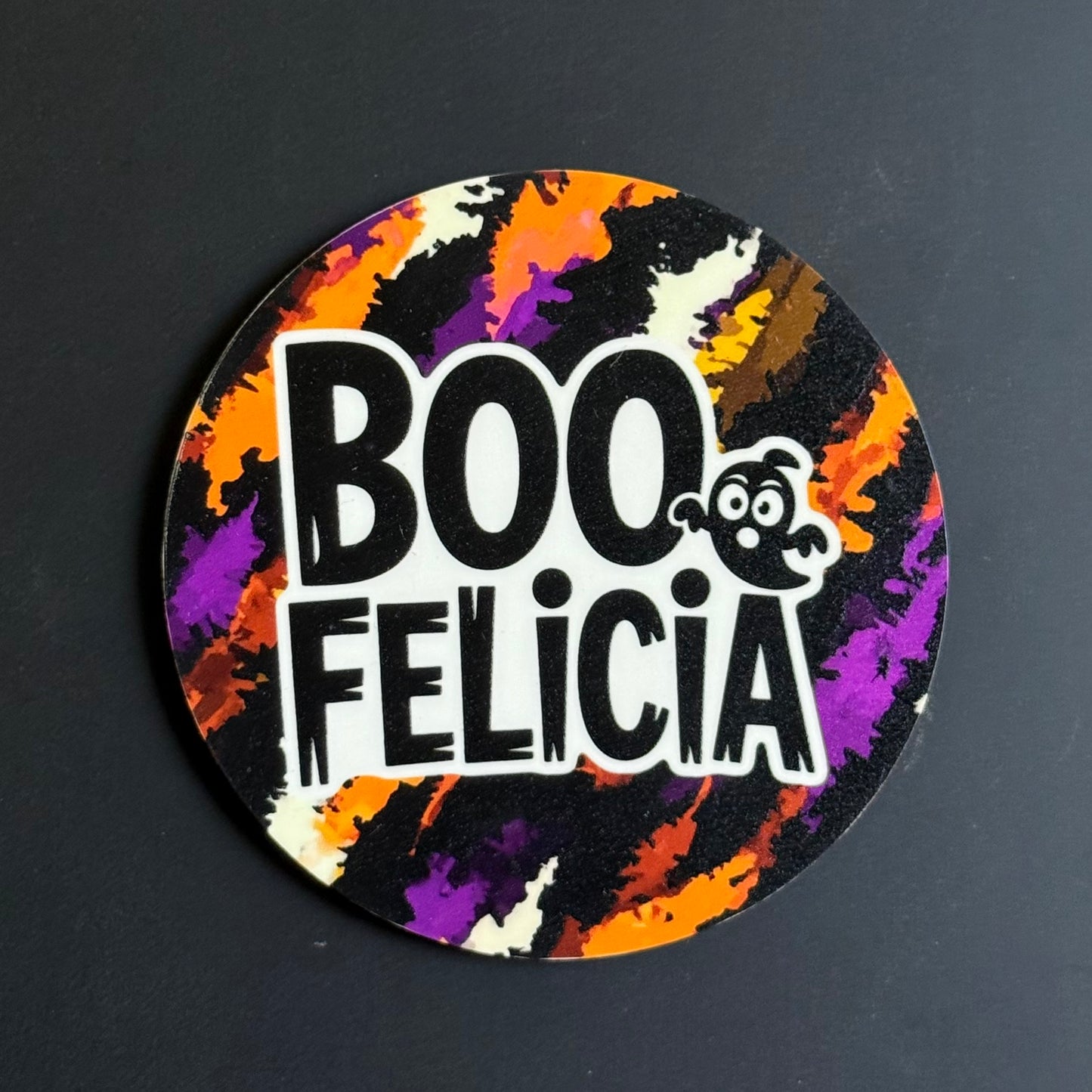 A circular sticker with a Halloween theme, featuring the text 'BOO FELICIA' in bold white letters with a small cartoon ghost. The background of the sticker has a colorful paint-splatter pattern in orange, purple, black, and cream.