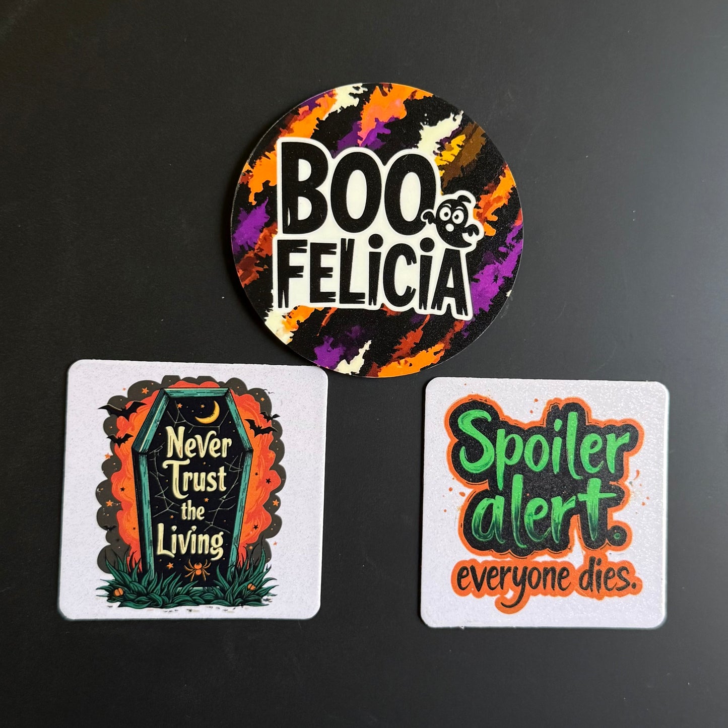 Three Halloween-themed printed items on a dark surface: a round sticker at the top reading “BOO FELICIA” with a colorful splatter background, and two square cards below with spooky quotes, one featuring a tombstone and the other reading “Spoiler alert. everyone dies.