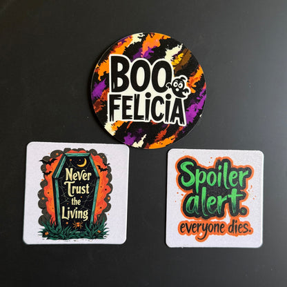 Three Halloween-themed printed items on a dark surface: a round sticker at the top reading “BOO FELICIA” with a colorful splatter background, and two square cards below with spooky quotes, one featuring a tombstone and the other reading “Spoiler alert. everyone dies.