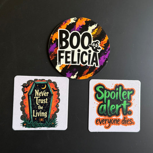 Three Halloween-themed printed items on a dark surface: a round sticker at the top reading “BOO FELICIA” with a colorful splatter background, and two square cards below with spooky quotes, one featuring a tombstone and the other reading “Spoiler alert. everyone dies.