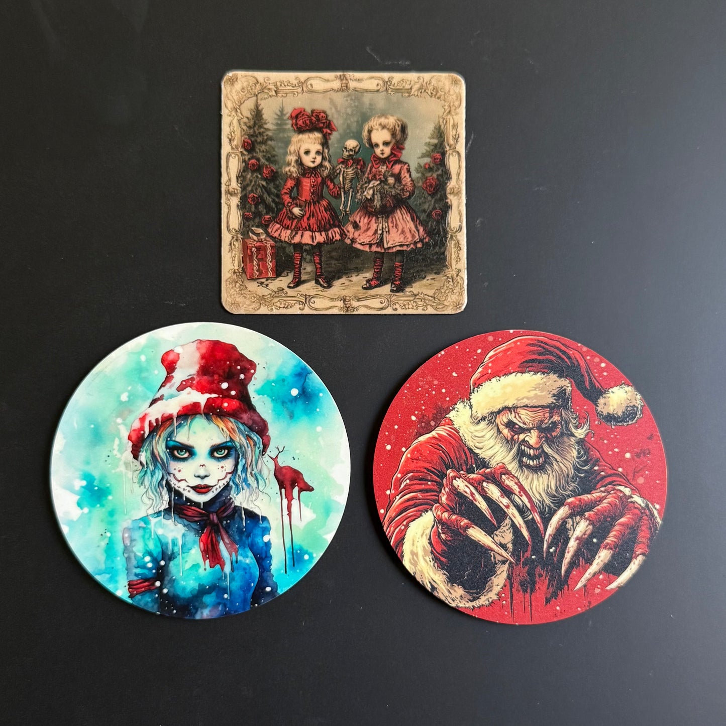 Three Halloween-themed art prints or coasters displayed on a dark surface: a square vintage-style print at the top showing two young girls in red dresses, and two circular prints below, one with a blue-toned zombie-like girl in a red hat and one with a red Santa Claus design.