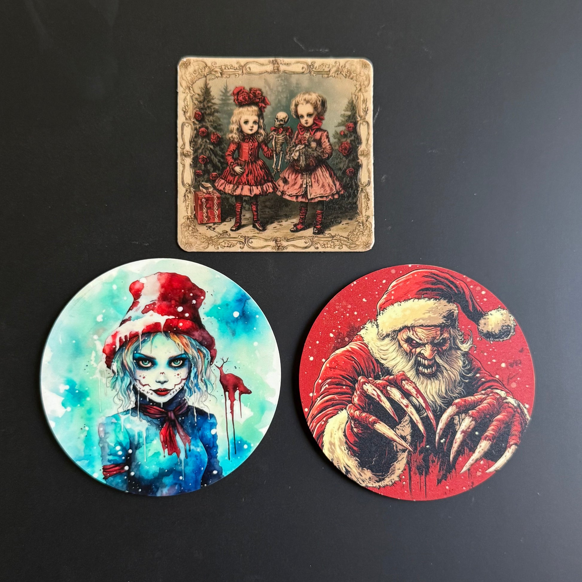 Three Halloween-themed art prints or coasters displayed on a dark surface: a square vintage-style print at the top showing two young girls in red dresses, and two circular prints below, one with a blue-toned zombie-like girl in a red hat and one with a red Santa Claus design.