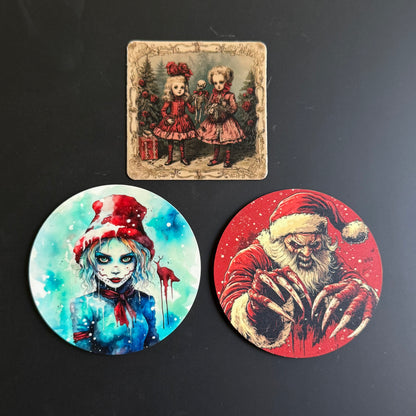 Three Halloween-themed art prints or coasters displayed on a dark surface: a square vintage-style print at the top showing two young girls in red dresses, and two circular prints below, one with a blue-toned zombie-like girl in a red hat and one with a red Santa Claus design.