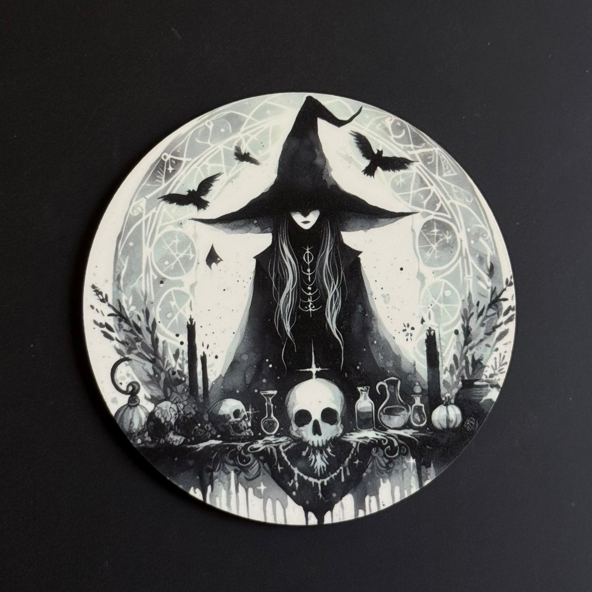 A circular coaster featuring a gothic witch illustration in black and white, depicting a witch in a tall hat with crows, a skull, potions, and candles, set against a dark background.
